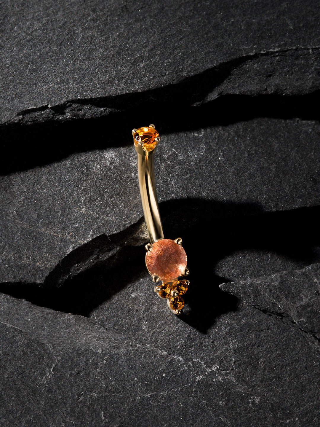 Gold belly button ring with orange gemstones on a dark stone surface