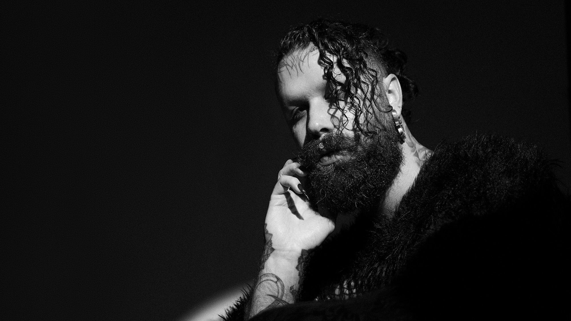 Close-up portrait of a man with a beard and intricate arm tattoos, part of a professional fashion photoshoot.